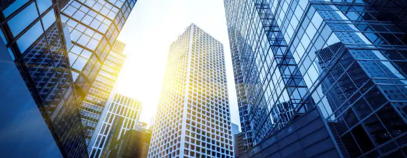 Tall modern skyscrapers with glass facades reflecting sunlight, viewed from below against a bright sky in an urban cityscape.