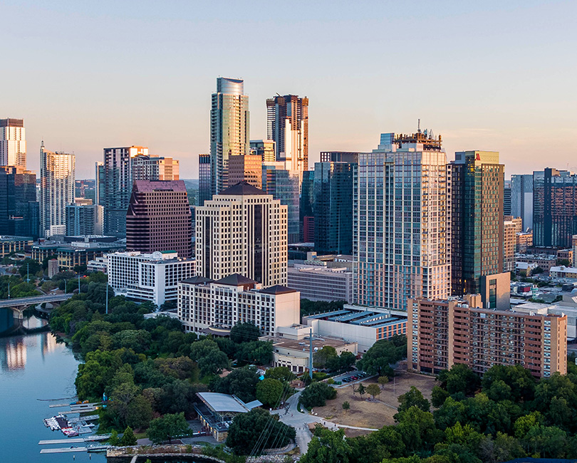 Aerial view of a city skyline with modern high-rise buildings, a river, and a bridge. Trees line the waterfront under a clear sky.