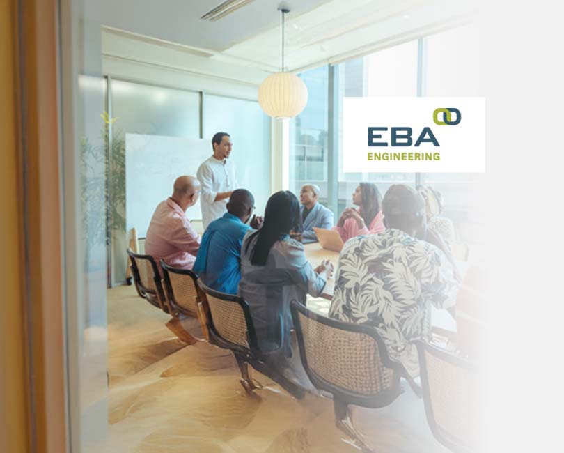A diverse group of people in a conference room sit around a table, engaged in business development planning. One person stands at the front, leading the meeting. Large windows reveal an urban landscape outside. The logo "EBA Engineering" is visible on the glass, highlighting its MBE certifications.