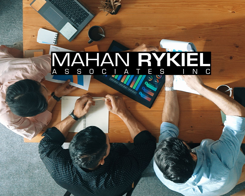 Three people sit around a wooden table adorned with a laptop, notebook, and documents, engaged in a lively leadership discussion. The overlay text reads "MAHAN RYKIEL ASSOCIATES INC.
