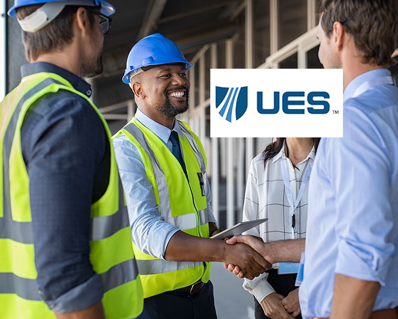A group at a construction site embodies a true success story. Two are in hard hats and neon vests, with one man in a blue hat shaking hands with a shirt-and-tie-clad colleague. A smiling woman stands beside them, embodying the spirit of business development in the unfinished building. UES logo prominently displayed.