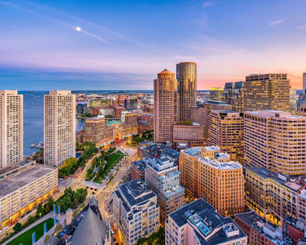 Aerial view of Boston, Massachusetts at sunset, showcasing a cityscape where modern skyscrapers meet historic buildings. The waterfront adds charm as the sky melds blue and orange hues, dotted with a few scattered clouds.