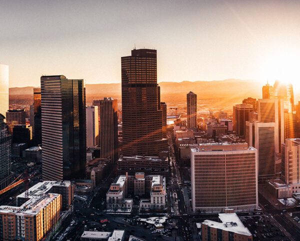 Aerial view of the Denver, Colorado skyline at sunset with tall skyscrapers casting long shadows. The sun is low on the horizon, creating a warm glow. Snow dusts the rooftops of several shorter buildings, and mountains rise majestically in the distance.