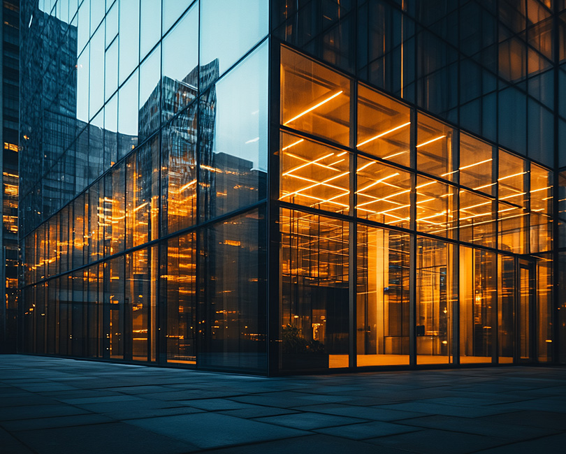 Glass building with illuminated orange interior, reflecting nearby skyscrapers. The scene is captured at dusk, highlighting the contrast between the bright lights and dark surroundings.