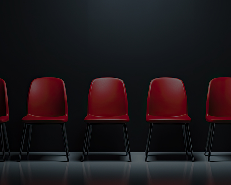 A row of seven red chairs with metal legs is evenly spaced against a dark wall, illuminated by overhead lighting, with reflections visible on the shiny floor.