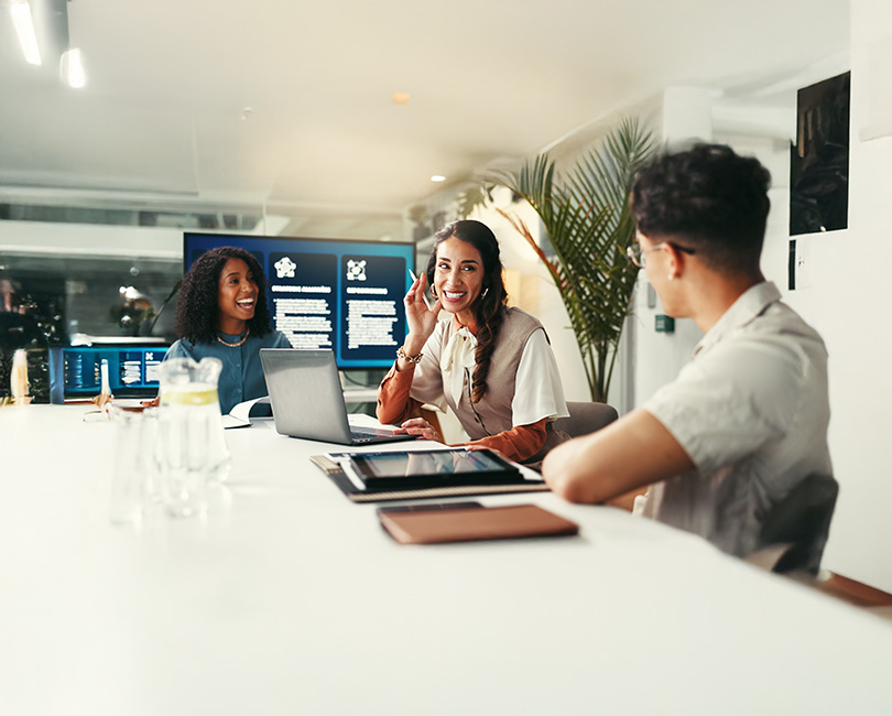 Three people sit at a conference table with laptops and notebooks, smiling and talking. Two large computer screens display graphs related to evergreen recruiting in the background, while a plant decorates the modern office space.