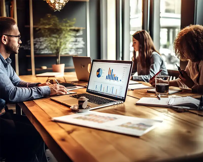 Two people work at a wooden table with laptops. One screen displays colorful charts and graphs. Papers, notebooks, a cup of coffee, and a glass of water are also on the table. Natural light fills the room.