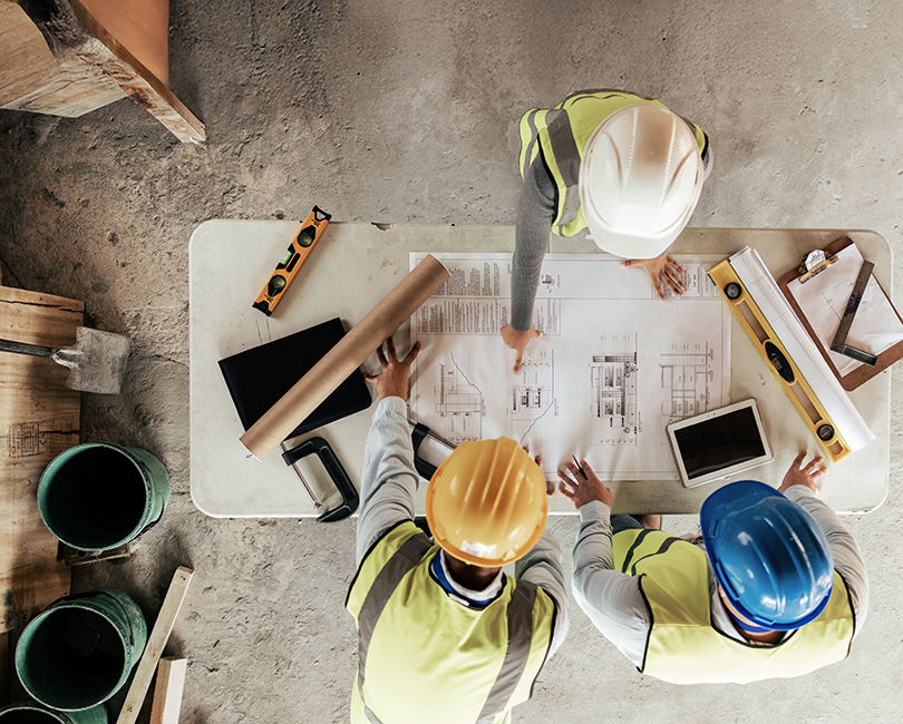 Two construction workers in safety vests and helmets review blueprints on a table surrounded by various building tools and documents. Text overlay reads, "Strategic Planning: Top Disruptions You Can't Ignore.