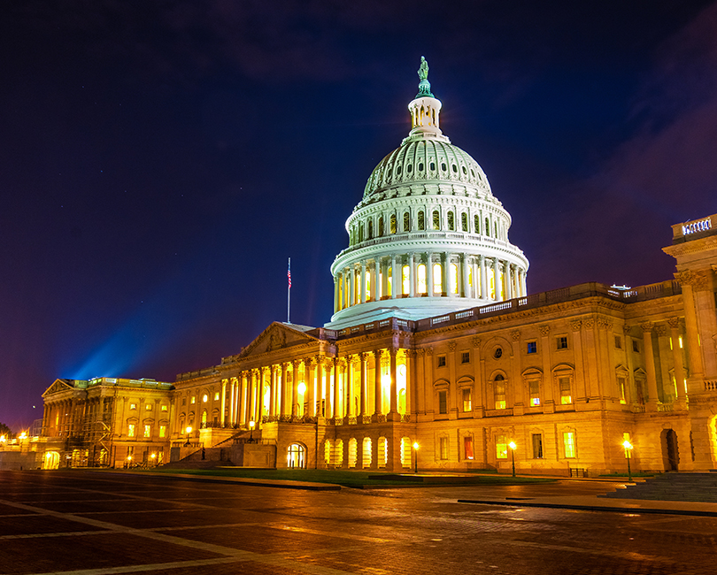 The U.S. Capitol building at night, brightly illuminated with golden lights, showcasing its iconic dome and neoclassical architecture against a dark blue sky.