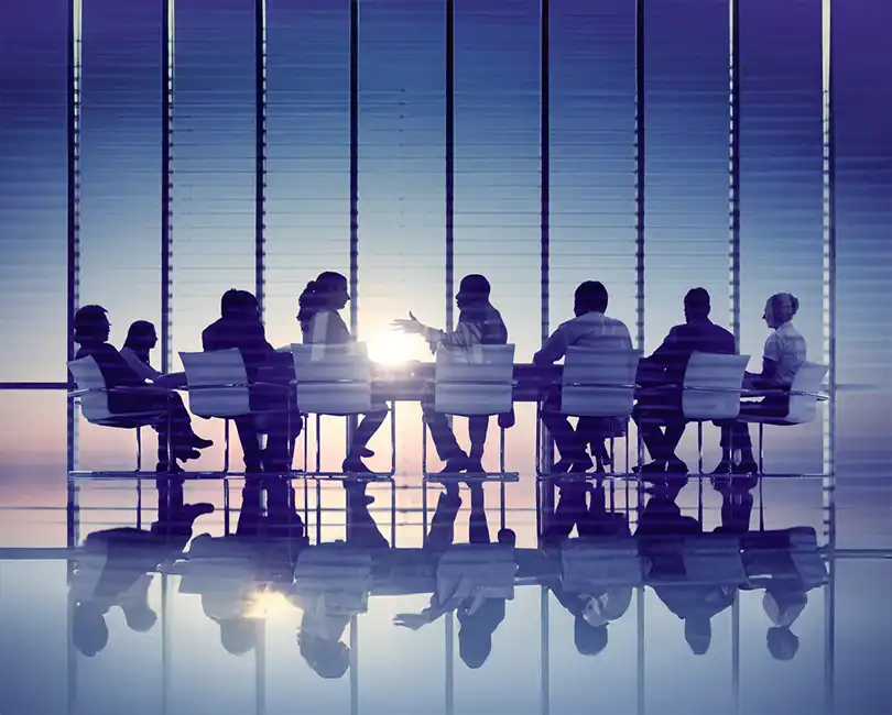 A group of people in business attire sit around a large conference table, engaged in discussion, with sunlight streaming through tall windows in the background.