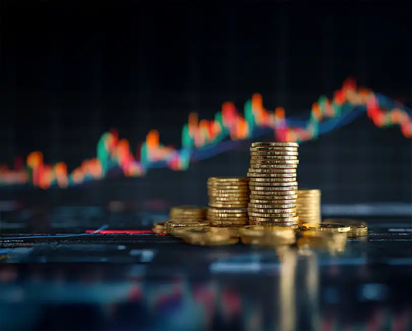 Stacks of gold coins are arranged in the foreground, with a blurred digital screen displaying colorful financial charts and graphs in the background, symbolizing investment and economic growth.