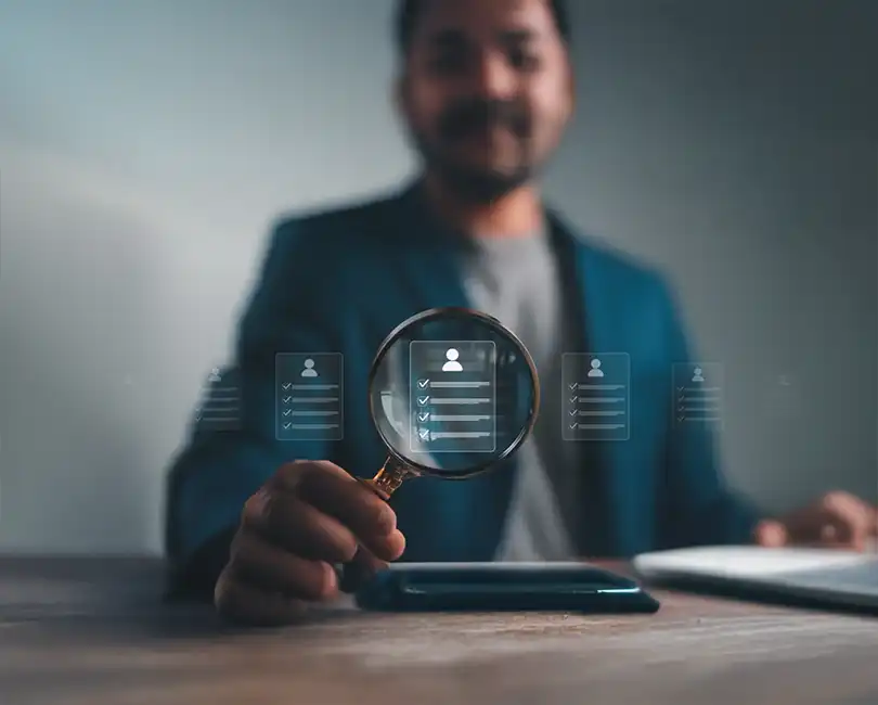 A person holding a magnifying glass focuses on a digital checklist icon with user profiles, suggesting the concept of candidate selection or recruitment. The person is blurred in the background, and the icons appear virtually.