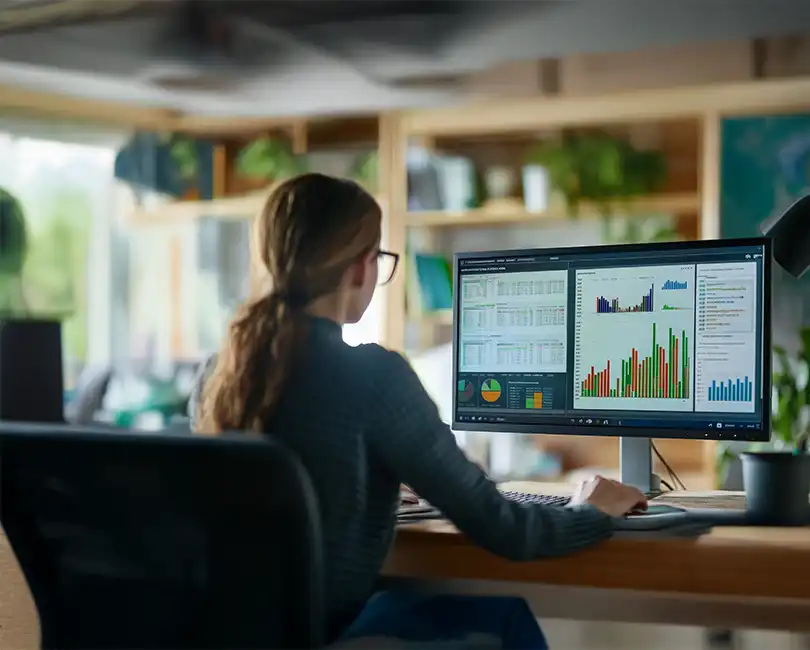 A woman sits at a desk in an office, analyzing colorful charts and graphs on a large computer monitor. Text on the image reads: "FAR Overhead Audits vs. Compilations: What DOT Contractors Need to Know