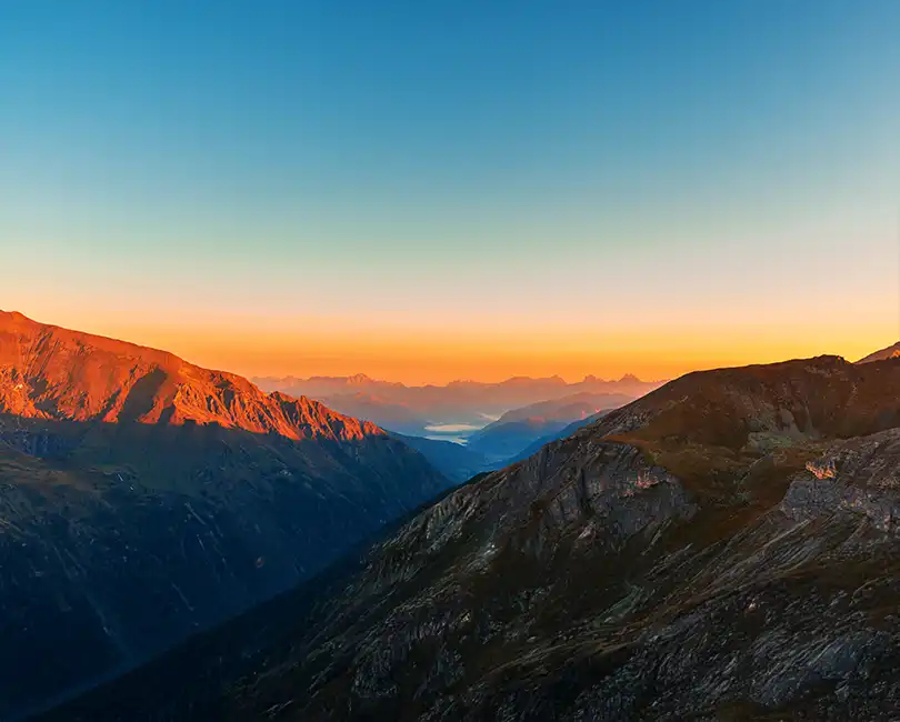 A scenic mountain landscape at sunset, with orange sunlight illuminating the slopes and distant peaks. The sky fades from deep blue at the top to warm orange near the horizon. A river or lake is visible in the far distance.