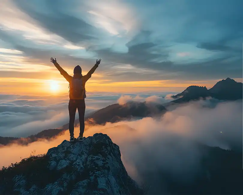 A person with a backpack stands on a rocky peak, arms raised toward a colorful sunrise above clouds and distant mountains, celebrating the view and achievement.