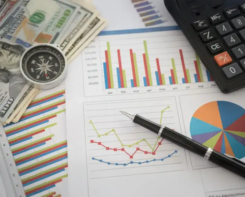 Charts, graphs, a calculator, a pen, a compass, and US dollar bills are spread out on a table, representing financial analysis and planning.