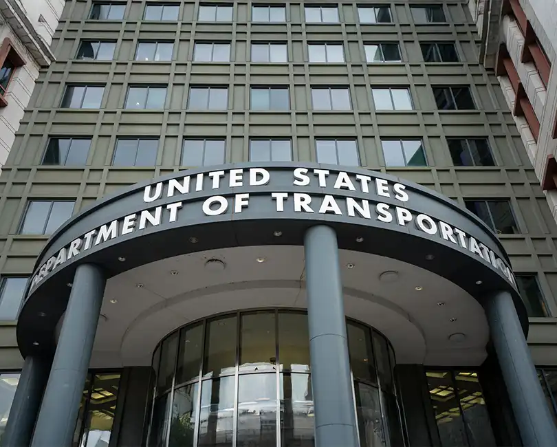 Main entrance of the United States Department of Transportation building, showing the curved awning with the department’s name and glass doors below, set against a tall office building facade.