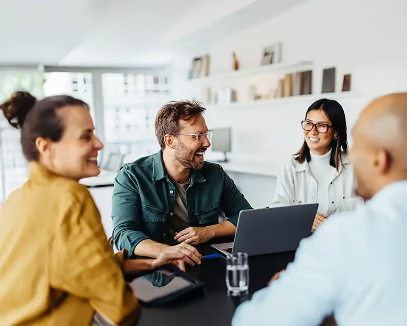 Four colleagues sit around a table, smiling and talking, with a laptop open in front of them. The text reads: “How to Give Constructive Feedback That Inspires Growth” by Kristi Weierbach, Ph.D., SHRM-SCP.