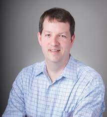 A professional headshot of Tom Godin, Sr. Director of Performance Consulting at Zweig Group, featuring a man with short brown hair, smiling and wearing a blue and white patterned button-down shirt against a neutral gray background.